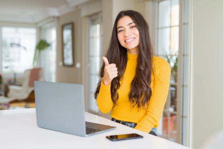 A smiling student sitting at a desk with a laptop and giving a thumbs-up gesture, showing confidence in her learning journey and academic success supported by Top Travel & Tourism Courses.